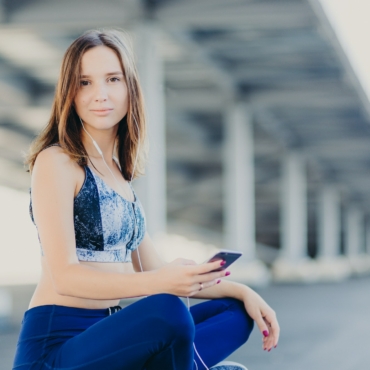 Attractive sporty woman sits crossed legs, holds modern cell phone, enjoys lively music in earphones