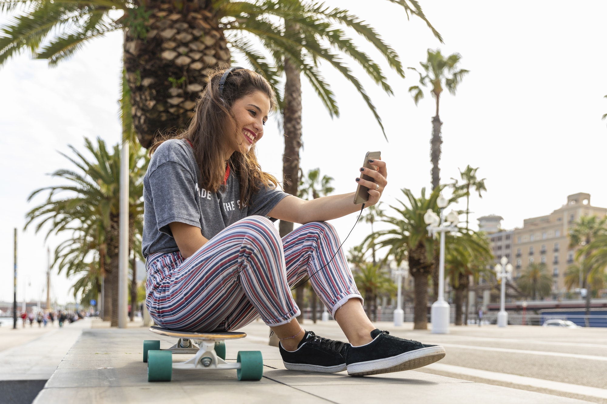 Happy teenage girl sitting on skateboard listening to music with headphones